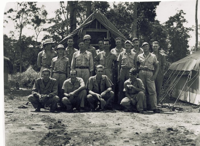 Sixteen men gathered in front of a tarpaulin-roofed hut with a wooden gable and screened window, jungle trees behind and a second tent pitched at the right, with another man cut off at the left edge of the frame. Four crouch in front, several wear sun helmets or soft caps, one has a pipe in his mouth, and one man's steel helmet carries a name painted across the front that is now only partly legible.