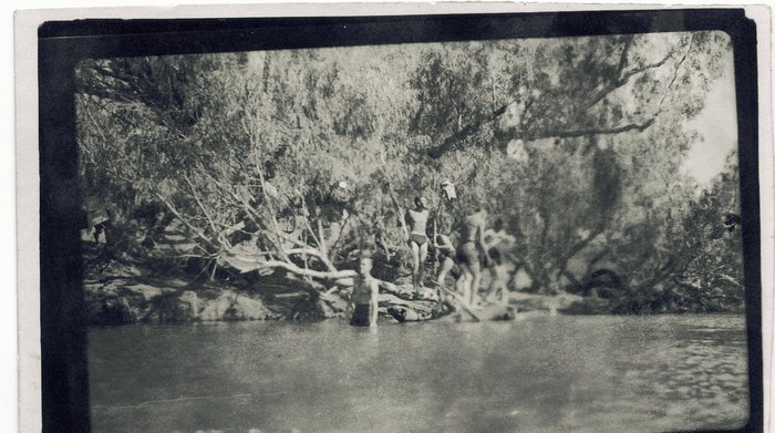 Half a dozen men at a swimming hole shaded by overhanging gum trees, one standing waist-deep in the water in a dark singlet while the others gather on the rocks and fallen limbs of the far bank. Clothing hangs drying from the branches above them.