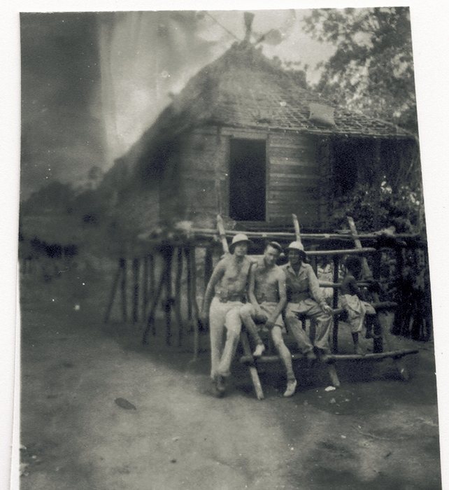 Three servicemen sit on a fence rail in front of a thatched hut, local children visible behind them.