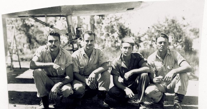 Four men crouch beneath an aircraft's undercarriage leg, the oleo strut and its linkage hanging from the flat underside of the airframe directly above their heads. Three wear light khaki shirts with rolled or cut-off trousers -- the man at left with a winged badge at the breast -- the fourth a darker work shirt and a knitted cap; two of them have moustaches.