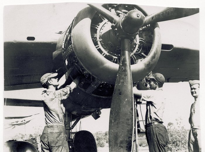 Mechanics working on an exposed twin-row radial engine, the propeller, nacelle and one mainwheel filling the frame. The blade carries its data stencil, legible when the print is turned upright: 