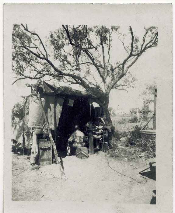 A man sits on a wooden crate at a bench inside the open end of a tent pitched under a spreading gum tree, its canvas fly stretched out over the entrance and propped on cut saplings. Packing cases stand at the doorway and a cable runs across the bare ground into the tent.