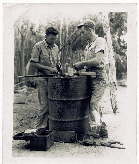 Two servicemen clean a rifle barrel on an oil-drum table.