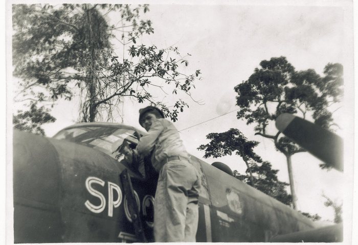 Spook II. A serviceman in light fatigues and a soft cap leans into her open cockpit, an access hatch swung down beside him, the first letters of her name -- <b>SPO</b> -- painted across the nose and hatch. Here they are outlined capitals with a drop shadow, where the frames that show the back of the name have it in solid white. An aerial wire runs from the canopy back along the fuselage, a small dorsal dome shows above it, and jungle trees rise behind.