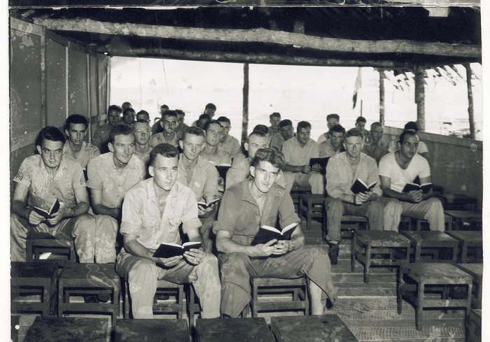 About twenty-five servicemen at a chapel service under a thatched roof.