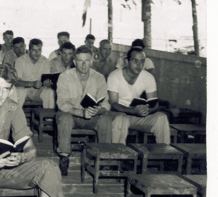 The same chapel service, closer in — Allen, in the white shirt, hymnal open beside another man still shirtless from the heat.