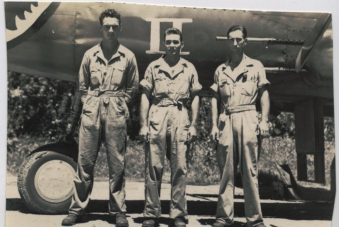 Crewmen standing at attention in front of a field-modified B-25 strafer. All that survives of her name is a Roman <b>II</b> in white serif capitals with a bar above and below; the word in front of it is cut off by the top edge of the print, and shark-mouth teeth show in the top left corner. She is Spook II. Another print in this collection shows the same nose from a little lower, with the word SPOOK standing above that same II; here the top edge of the paper has cut it off. This caption used to call her 