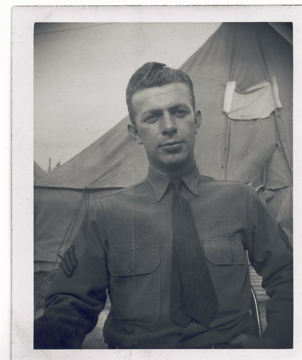 Waist-up portrait of a sergeant in a wool service shirt and tie, three chevrons on his sleeve, seated in front of a pyramidal tent with a pale towel hung over its front.