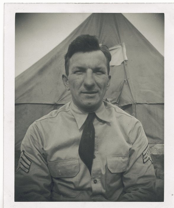 A sergeant, chevrons visible, stands before a tent.