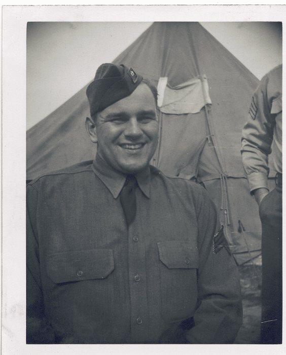 A grinning serviceman in a wool shirt and tie, a small metal insignia pinned to the curtain of his garrison cap, photographed in front of a pyramidal tent; a second man stands at the right edge of the frame with chevrons on his sleeve.