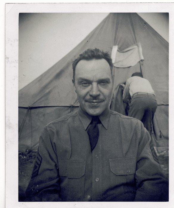 A moustached serviceman in a wool shirt and tie, chevrons just visible at the cut edge of his sleeve, looks into the camera while behind him another man in a light shirt works at the open flap of a pyramidal tent.