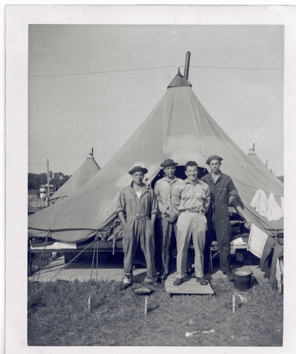 Allen is one of four men outside a pyramidal tent with a stovepipe through its peak: two in one-piece work coveralls and soft hats, one in shirt and trousers with a pipe in his mouth, and one bare-headed in shirtsleeves standing on a wooden duckboard. Folding cots, hanging towels, a pan and a bucket stand about the tent, with more tents and a pole line beyond.
