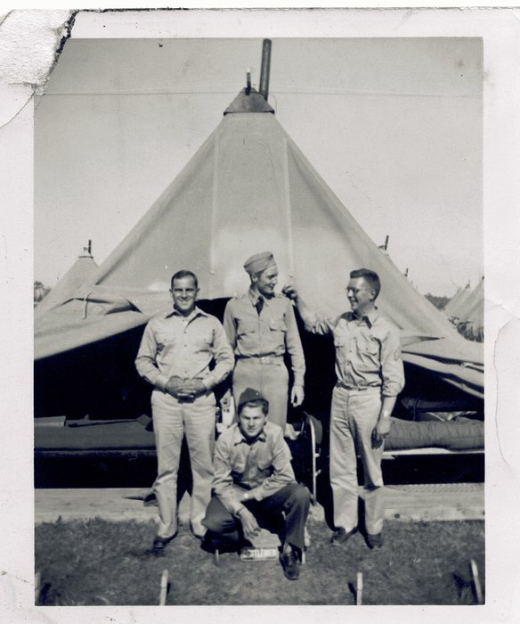 Four soldiers, one crouching, smile for a group photo among the tents.