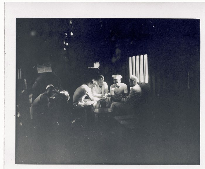 Six men in a dark room, several in undershirts and light fatigue caps, their hands gathered at the centre of the group and a metal basin on the table in front of them. Bare bulbs hang from the ceiling, a printed sheet is tacked to the wall at the left, and the only light comes through a slatted window at the right.