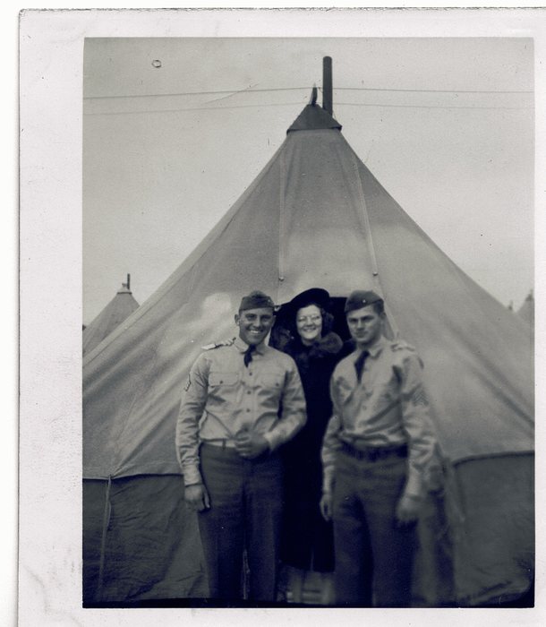 Allen is one of two servicemen in khaki shirts and ties with garrison caps standing either side of a woman in a dark fur-collared coat and hat at the entrance of a pyramidal tent, the stovepipe rising behind them and further tents on both sides.