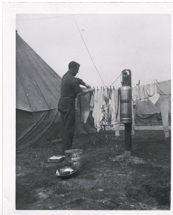 Allen, in wool shirt and trousers, pegs washing to a line strung between tents, a soda-acid fire extinguisher hanging from a board on a post beside him. On the ground are a washbasin, a flat cardboard box, and a galvanized bucket stencilled "NO. 3" and hand-lettered below with "SGT" and a name ending "...ALD".