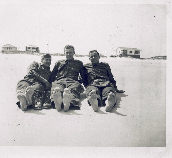 Allen and two other soldiers relax on a beach, wood-frame cottages behind them.