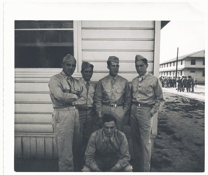 Five men in cotton service shirts, ties and garrison caps at the corner of a wooden barracks building, Allen at the left &mdash; four standing and one seated on the ground in front of them. A second two-storey barracks stands beyond, with a group of roughly twenty more men gathered along the road behind.
