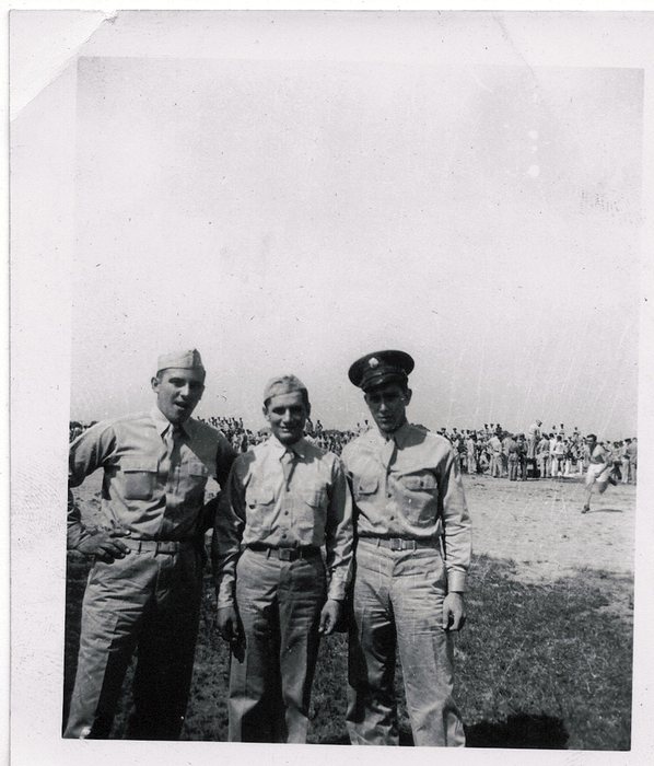 Three men in cotton service shirts and ties in an open field, Allen at the left: two in garrison caps and the third in a visored service cap with a cap badge. A long line of men is massed across the background, and at the right a man in shorts and a light shirt runs past.