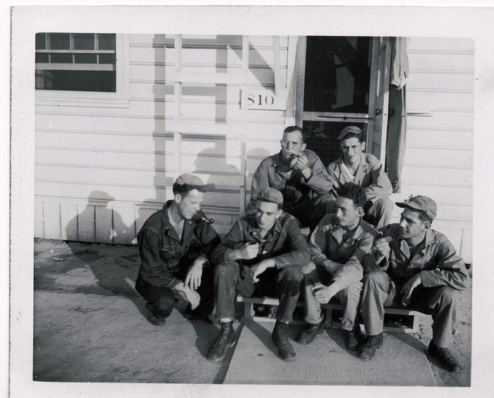 Six soldiers, several smoking pipes, relax on the steps of barracks 810.