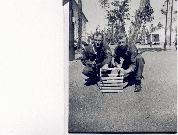Allen at the left, with another man in fatigue coveralls, crouching on an unpaved camp road holding up a home-made crate of light wooden slats fitted with a carrying handle, something dark visible inside it; Allen wears chevrons on his sleeve and each of them holds a tool. Wooden camp buildings, tall pines, a parked 1930s sedan and a distant figure near the far building fill the background.