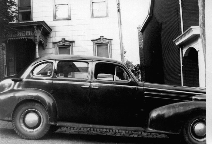 A 1930s sedan parked on a street of Victorian rowhouses.