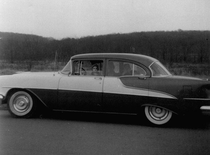 A two-tone four-door sedan with whitewall tyres on a paved road below a wooded ridge of bare trees. John Sweigert, Allen's younger son, looks out through the front side window, with two further figures indistinct in the back seat.