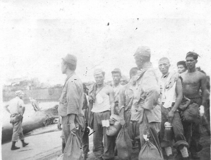 A group of captured Japanese soldiers, eight or nine visible, stands on a shoreline track with their belongings tied up in cloth bundles and a canteen in one man's hand; some are in uniform shirts and field caps, others stripped to undershirts or bare to the waist. A further man in a light shirt and soft cloth cap walks along the track behind them, with water and a far shore beyond.