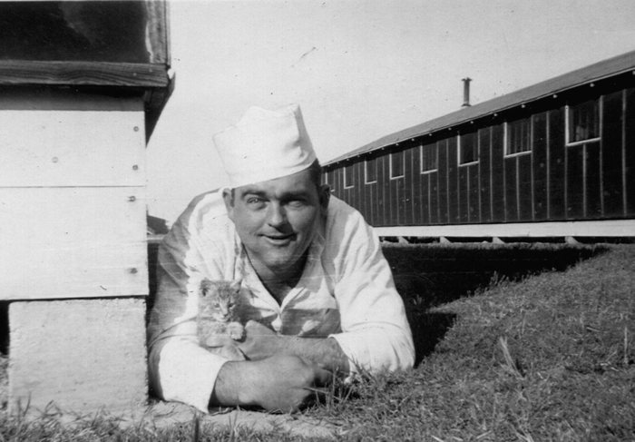 A man in a cook's white jacket and white cap lies on the grass beside a wooden building, holding a tabby kitten against his chest. A long dark barracks with a row of windows and a stovepipe on the roof runs away behind him.