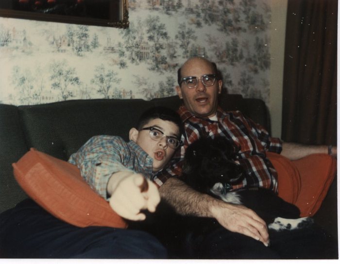 A colour snapshot of two people on a dark green sofa banked with orange cushions: Allen in heavy-framed glasses and a red plaid shirt, and his son John in similar glasses and a blue plaid shirt, with a black-and-white dog lying between them. One of them reaches a hand out toward the camera; the wall behind carries a printed scenic wallpaper and the corner of a gilt picture frame shows at the top left.