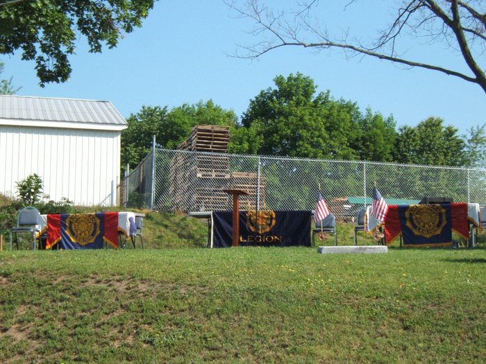 Folding chairs and three banner-draped tables stand ready on the grass before a service begins, the centre drape carrying the American Legion emblem and the word "LEGION", with a plain wooden lectern beside it. Two small United States flags in bronze grave-marker holders and a low white marker stone are set out at right, with a metal-sided building, a chain-link fence and stacked wooden pallets behind.