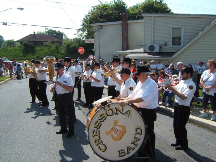 A community band plays in the street, the bass drum lettered "CRESSONA BAND" around a gold lyre; some twenty musicians in white shirts, black trousers and dark caps work through clarinets, trumpets, saxophones and a tuba. Spectators line the kerb, a "STOP" sign stands at the corner, and a small sign on the building behind reads "AMERICAN LEGION".