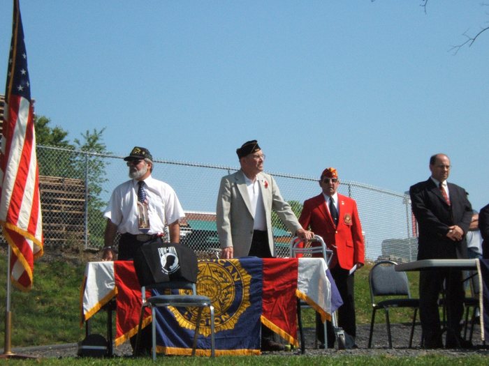 Four men stand behind a table draped with the American Legion emblem banner. Allen is second from the left, in a garrison cap and tan jacket with his hand on his walking frame and a red crepe poppy at his lapel; at the end of the row, one in a garrison cap, white shirt and dark tie, one in a red blazer and cap, and one in a dark suit. A black POW-MIA cloth is laid over the chair in front, and a United States flag on a stand rises at left.