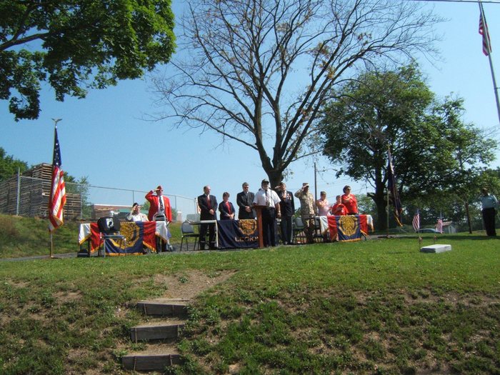 A wide view of the service: a man in a garrison cap stands at the lectern in front of a table draped with the word "LEGION", with about a dozen people ranged behind banner-draped tables, four of them saluting and one in desert-pattern camouflage. A United States flag stands unattended in a floor stand at left and furled flags on planted poles at right; a white marker stone with two small flags sits on the grass, and timber steps climb through the foreground.
