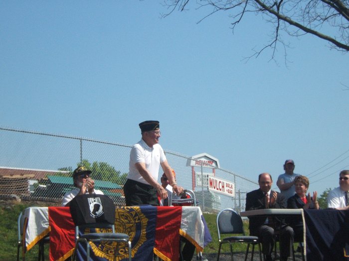 Allen, in an American Legion garrison cap and white polo shirt, stands behind the emblem-draped table with his hand on his walking frame, mouth open in a laugh, while those seated around him applaud; a POW-MIA cloth hangs over the chair beside him. A commercial sign beyond the chain-link fence reads "MULCH BULK + BAG".