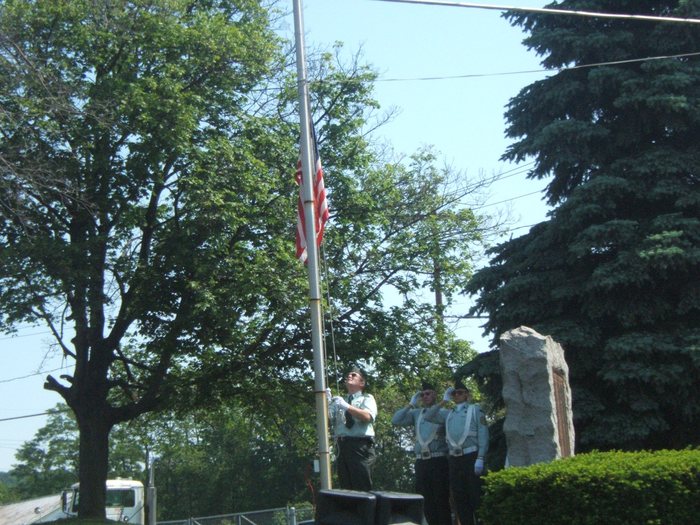 A man in a light uniform shirt and sunglasses works the halyard with the flag hanging partway up the pole, while two colour-guard members in pale blue shirts, white belts and white gloves stand at the salute. A rough-cut granite memorial stone stands at right against the evergreens.