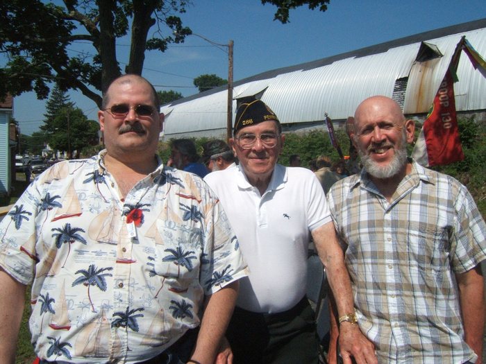 Allen with both his sons, shoulder to shoulder beside a corrugated-metal building &mdash; John Sweigert at the left, Allen in the centre, Allen Sweigert, Jr. at the right. His cap is lettered "286" and "PENN..." and seated with a chair or wheeled walker behind him. The man at left has a red crepe poppy pinned to his shirt, and furled parade banners on staffs stand against the wall behind, the red one partly readable as "...ROOP 130".