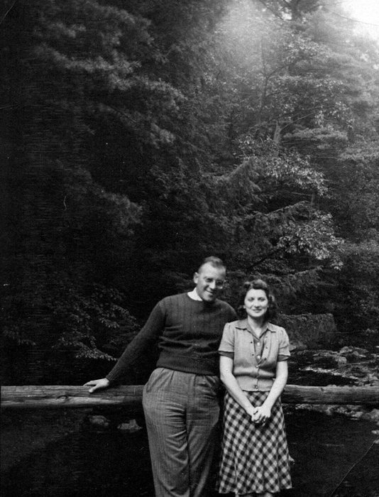 Allen, in a crew-neck sweater and wire-rimmed glasses, leaning on a rustic log rail beside Catherine in a short-sleeved buttoned blouse and a checked skirt, her hands clasped in front of her. Dense conifers and hardwoods cover the slope behind them.