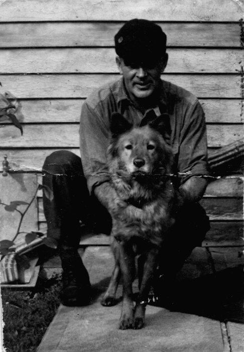 Earl Joshua Sweigert, Allen&rsquo;s father, in a dark flat cap, a work shirt with the sleeves rolled back, dark trousers and high boots, sitting against the clapboard wall of a house and holding a thick-coated dog steady between his knees. A creeper climbs the wall at the left.
