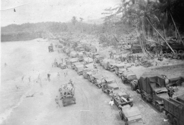 Seen from above, a double column of trucks, trailers and jeeps stands nose to tail along the edge of the palms at a landing beach, with men moving across the open sand and small craft drawn up at the waterline in the distance. A large pale circular object -- a spare wheel or a coil -- rides on top of the load of the nearest truck.