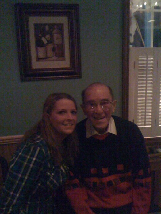 Allen sits cheek to cheek with his granddaughter Jessica in a restaurant, in a red and navy patterned sweater and heavy-framed glasses; she is in a green plaid shirt. A framed still life of wine bottles hangs on the wall above them and white louvered shutters stand at the right.