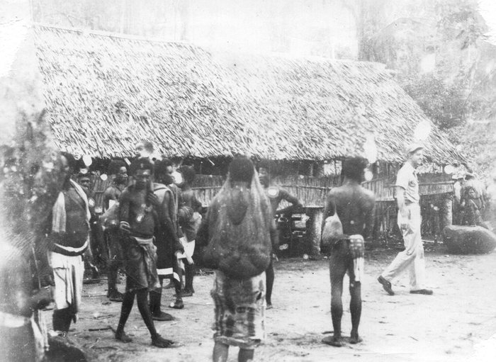 A gathering outside a thatched longhouse; a serviceman walks through, one villager carrying a woven net bag.