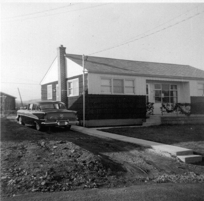A single-storey house seen from the driveway, with a brick chimney, two-tone siding, a shingled roof and a decorated front-porch railing. A two-tone sedan is parked on the drive, the concrete walk and steps are newly poured, and mounded bare earth fills the foreground.