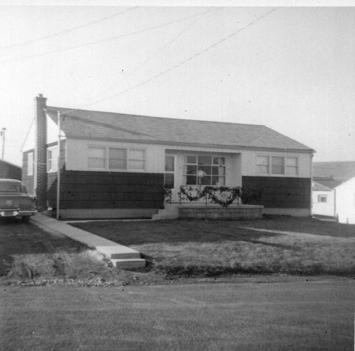 The front of the same low house from across the street, evergreen garland swagged along the porch railing with a pale ornament in each loop and the front of a car cut off at the left edge. The new concrete walk runs down to two freshly poured steps set in raw earth and gravel at the street.