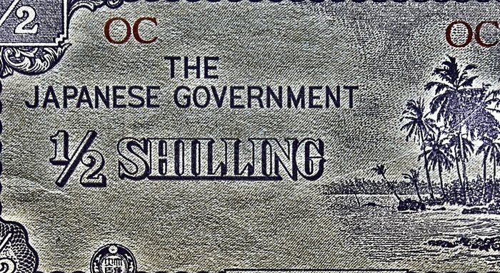 A tight crop of one side of the Japanese Government occupation half-shilling note for Oceania, showing "THE JAPANESE GOVERNMENT" over "1/2 SHILLING", palms and a shoreline at right, the corner fraction "1/2" clipped by the crop at upper left, and the red "OC" overprint twice along the top.