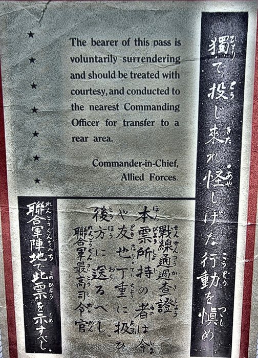 The English side of the same Allied safe-conduct leaflet, printed "The bearer of this pass is voluntarily surrendering and should be treated with courtesy, and conducted to the nearest Commanding Officer for transfer to a rear area." and signed "Commander-in-Chief, Allied Forces.", with a column of seven stars down the left margin and Japanese vertical panels at right and lower left; this is word for word the text Allen reads aloud on camera.