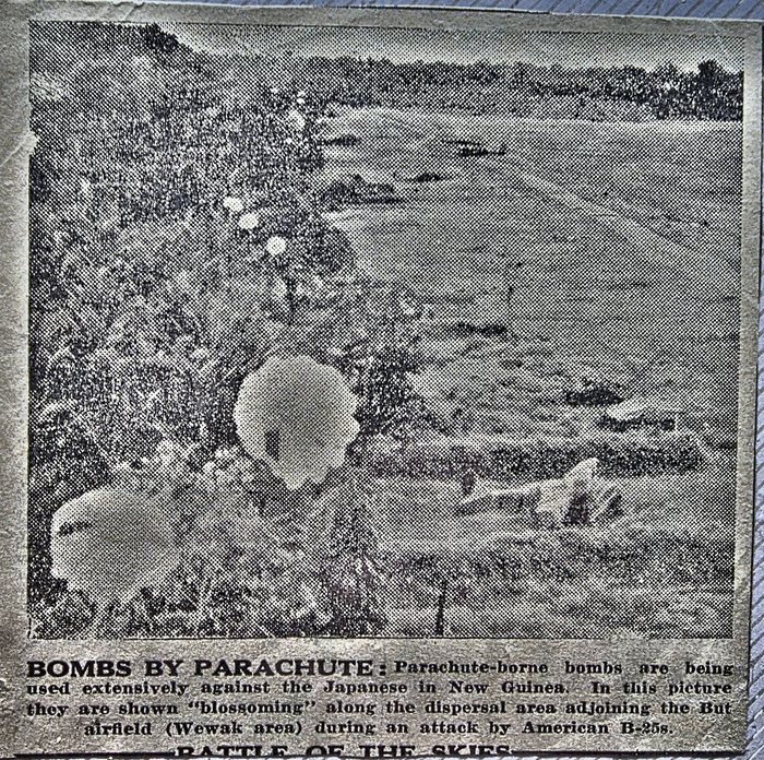 A newspaper clipping he kept: an aerial photograph in which pale parachutes hang scattered over scrub and open ground, with the printed caption "BOMBS BY PARACHUTE: Parachute-borne bombs are being used extensively against the Japanese in New Guinea. In this picture they are shown "blossoming" along the dispersal area adjoining the But airfield (Wewak area) during an attack by American B-25s." A second headline, "BATTLE OF THE SKIES", is cut off at the bottom edge. Credit: the newspaper is not named on the cutting; from his own collection.