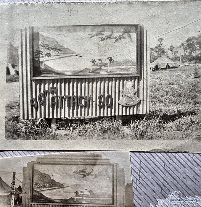 A hand-made squadron signboard of corrugated iron, its raised letters reading "90TH ATTACK SQ" below a painted panel of a palm-fringed bay with aircraft flying over it, and a scythe-bearing figure on a shield at the right end. Pyramidal tents stand in the grass behind the board, and the edge of a second print of the same sign shows along the bottom of the frame.