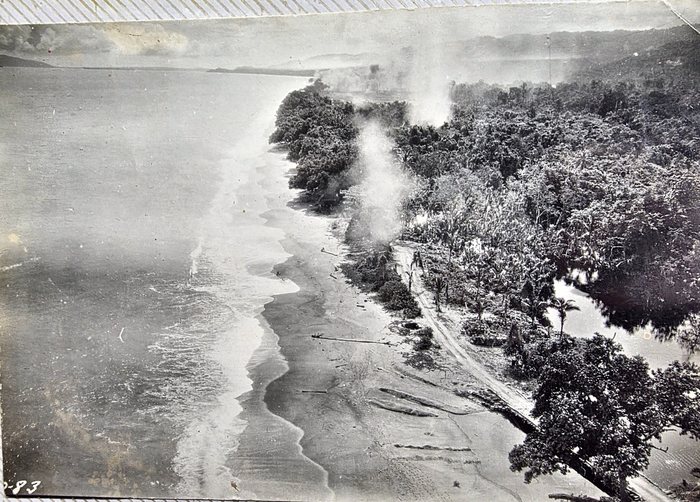 A low oblique aerial photograph looking down on a jungle shoreline, with columns of pale smoke rising from among the trees just inland of the beach and a track running along behind the sand. A number is inked on the print at lower left, partly cropped, reading "-83".