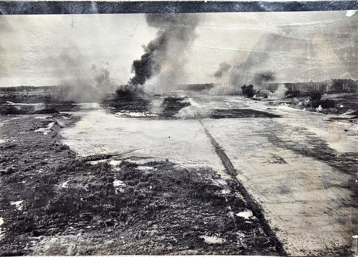 A low-level aerial photograph taken along a cleared airstrip, its matted surface and a long strip of runway matting running away from the camera. A tall column of black smoke and several lighter bursts rise from the far end of the strip and from the treeline beyond it.
